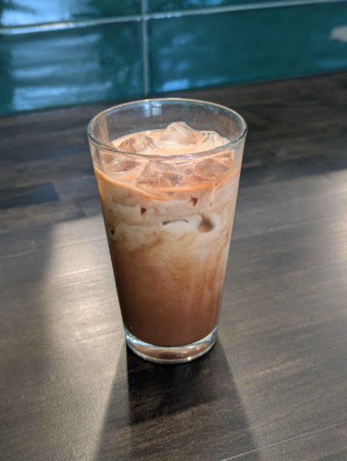 Photo of an iced mocha in a pint sized glass on a dark brown wooden countertop.