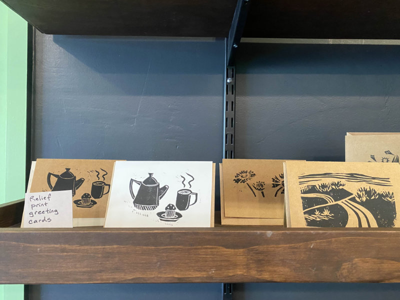 Zoomed in photo of a portion of the wooden merchandise shelf. Showcases a variety of differently designed greeting cards printed in black ink.