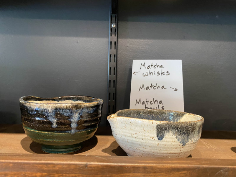 Zoomed in photo of a portion of the wooden merchandise shelf. Showcases 2 handmade matcha bowls. The bowl on the left is black on top and green at the bottom. The bowl on the right is cream colored with some gray spots. Both bowls have spouts for pouring.