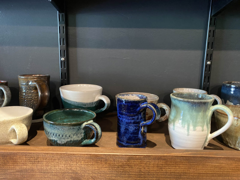 Zoomed in photo of a portion of the wooden merchandise shelf. Showcases handmade pottery, specifically various sized and colored mugs.