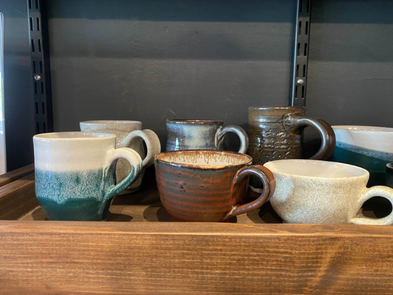 Zoomed in photo of a portion of the wooden merchandise shelf. Showcases handmade pottery, specifically various sized and colored mugs.