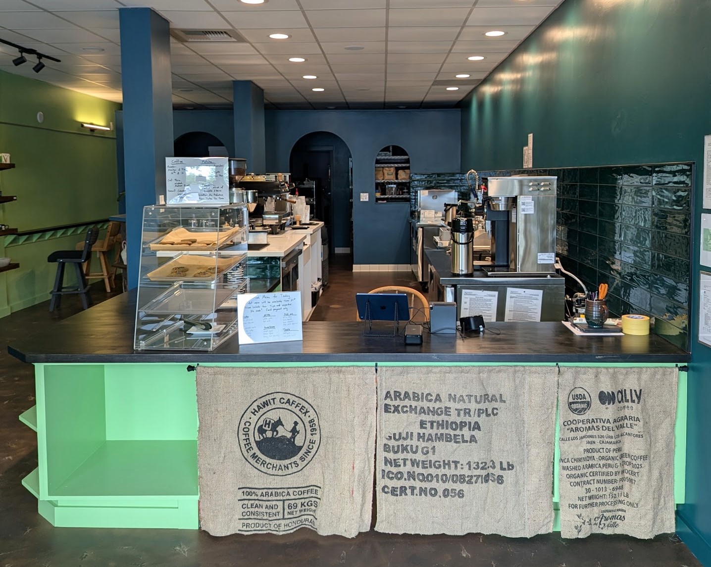Photo of the Fair Grounds building interior. The walls are painted green, blue, and teal and the photo is taken in front of the green store counter, with the menu and pastry case in view.