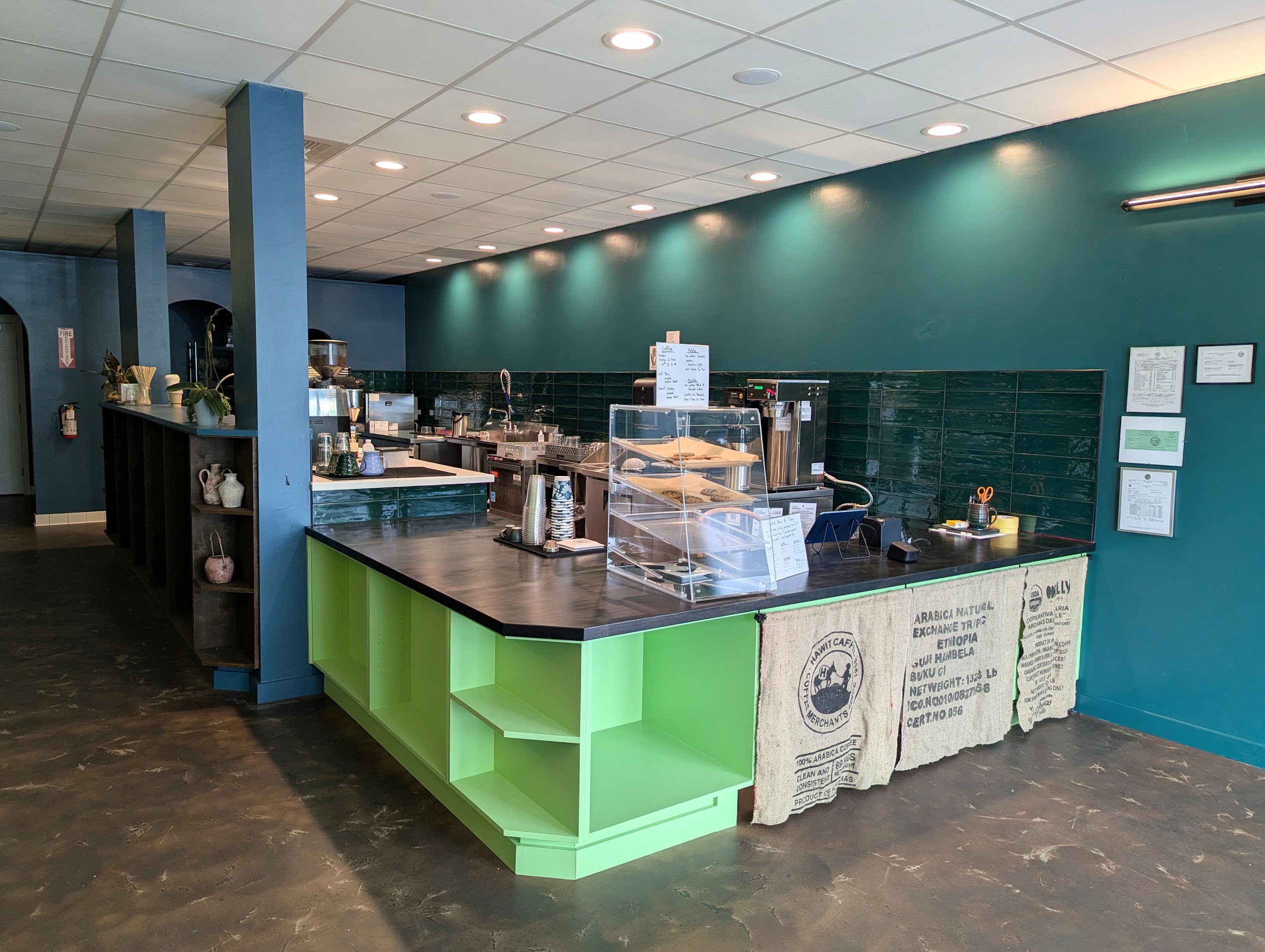 Photo of the Fair Grounds building interior, facing the kitchen. Shows the green counter with a menu and pastry case on top, and white kitchen countertops with espresso and drip machines and teal tiled backsplash.