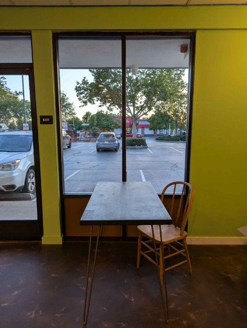 Photo of the Fair Grounds building interior, facing the shop front. Shows a tall window facing the parking lot with a dark brown wooden table and light brown wooden chair in front.