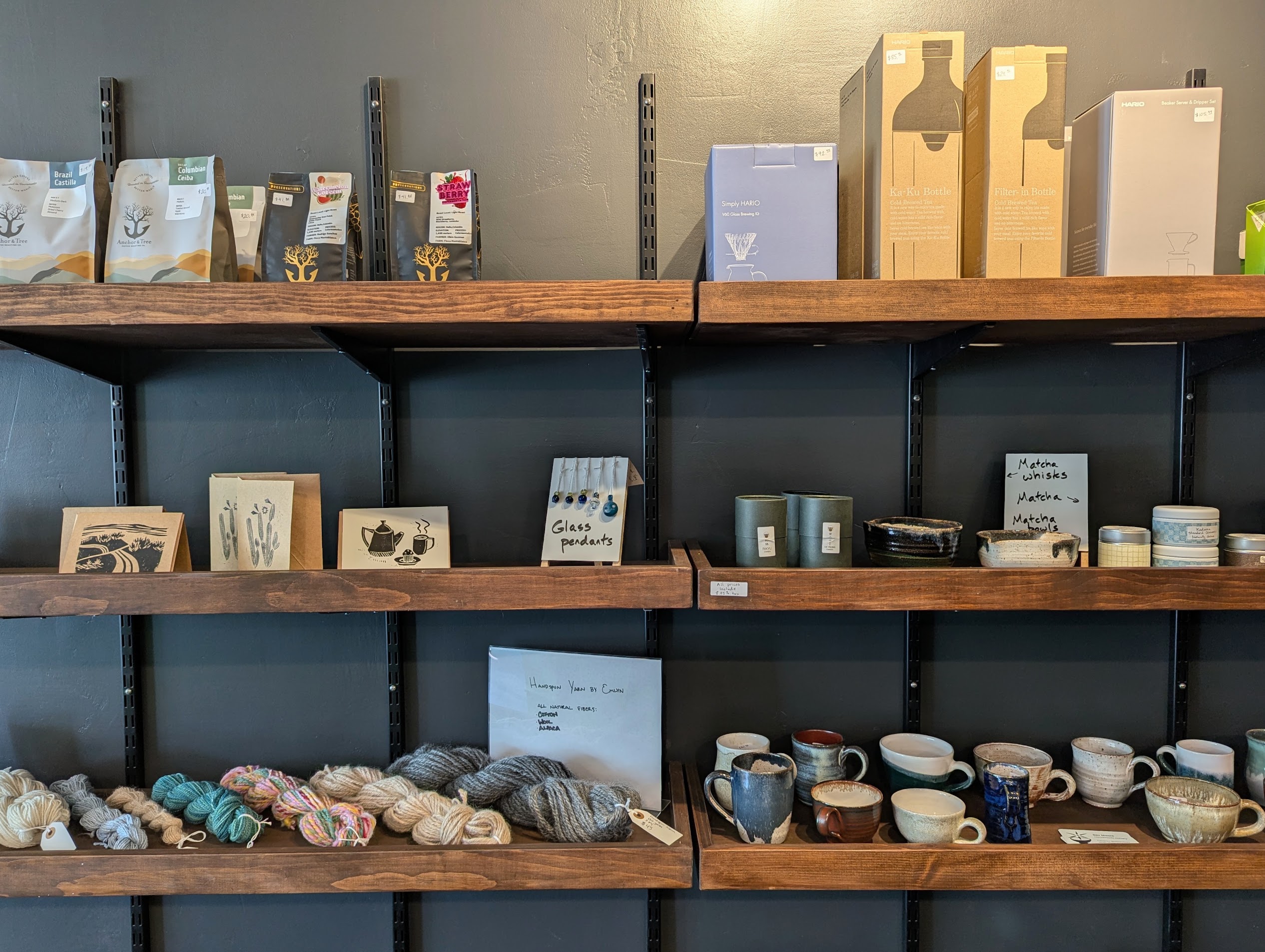 Photo of the Fair Grounds merchandise shelf. Shows three rows of wooden wall shelves. On the top shelf are bags of coffee beans, and tea bottles and filters. On the middle shelf are greeting cards, glass pendants, matcha, matcha bowls, and matcha whisks. The bottom shelf has various colored yarns and handmade pottery.